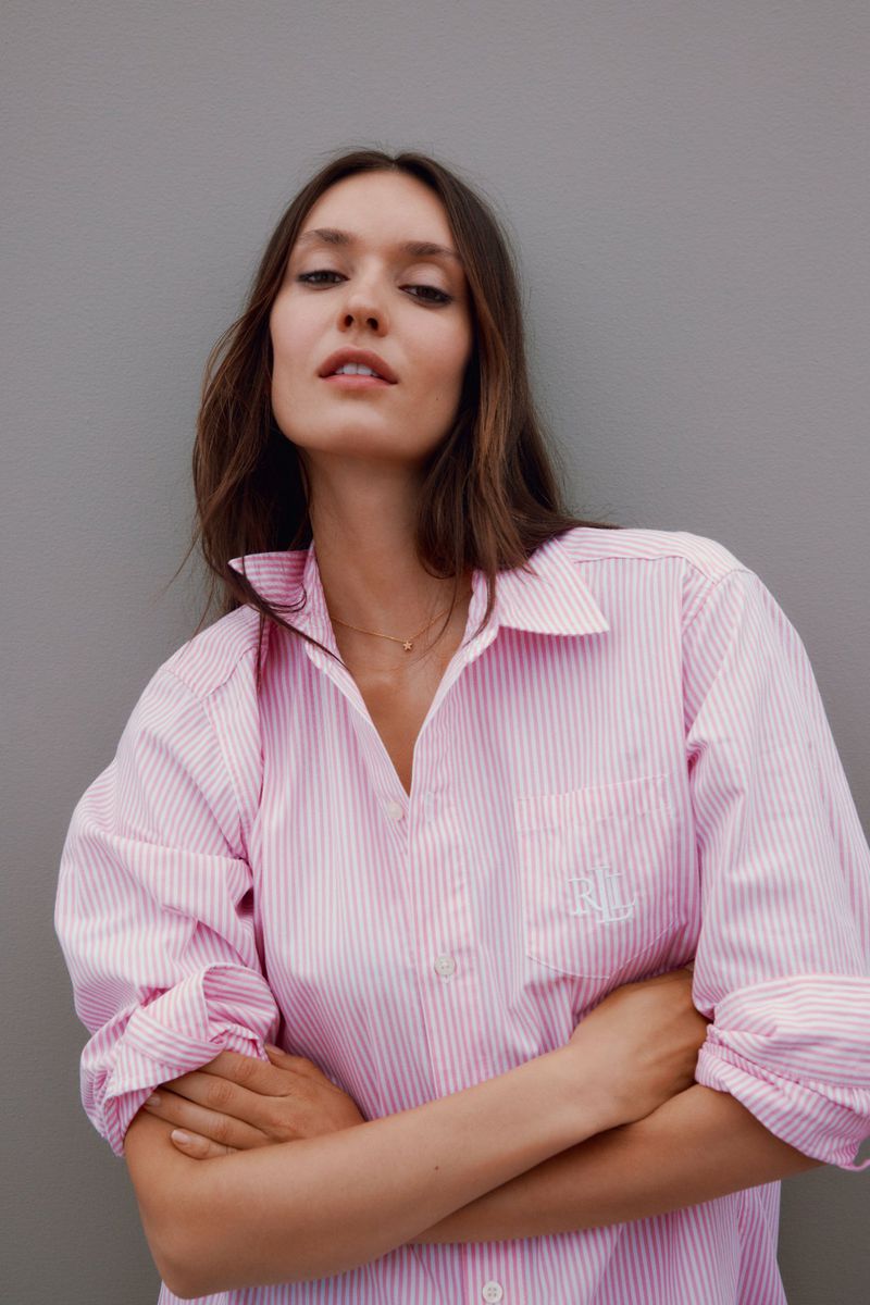 A young woman poses indoors wearing a pink-and-white striped shirt with her arms crossed, against a solid gray background.