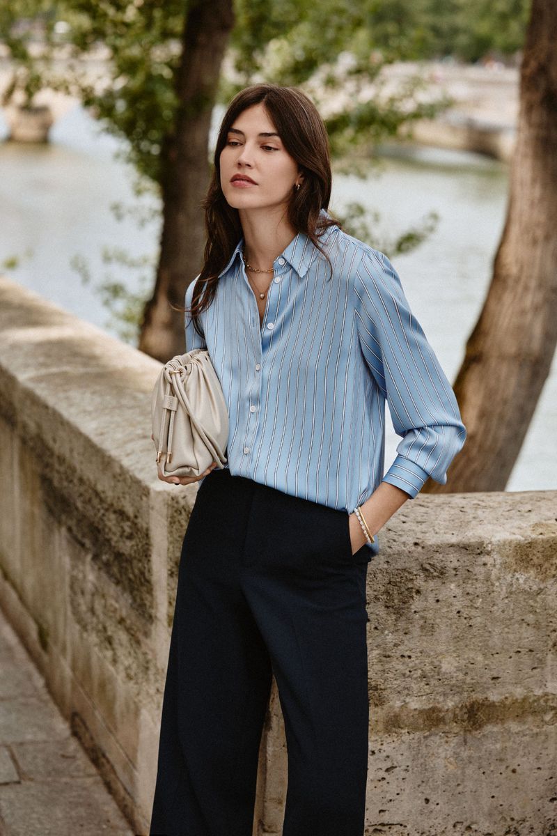 Marilhea Peillard wearing a light blue button-up shirt and black wide-leg trousers leaning against an outdoor stone wall.