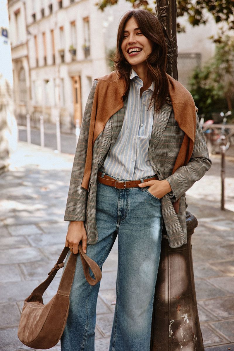 A smiling woman in a blazer and jeans walks down a Parisian street with a leather shoulder bag, posing near a lamp post.