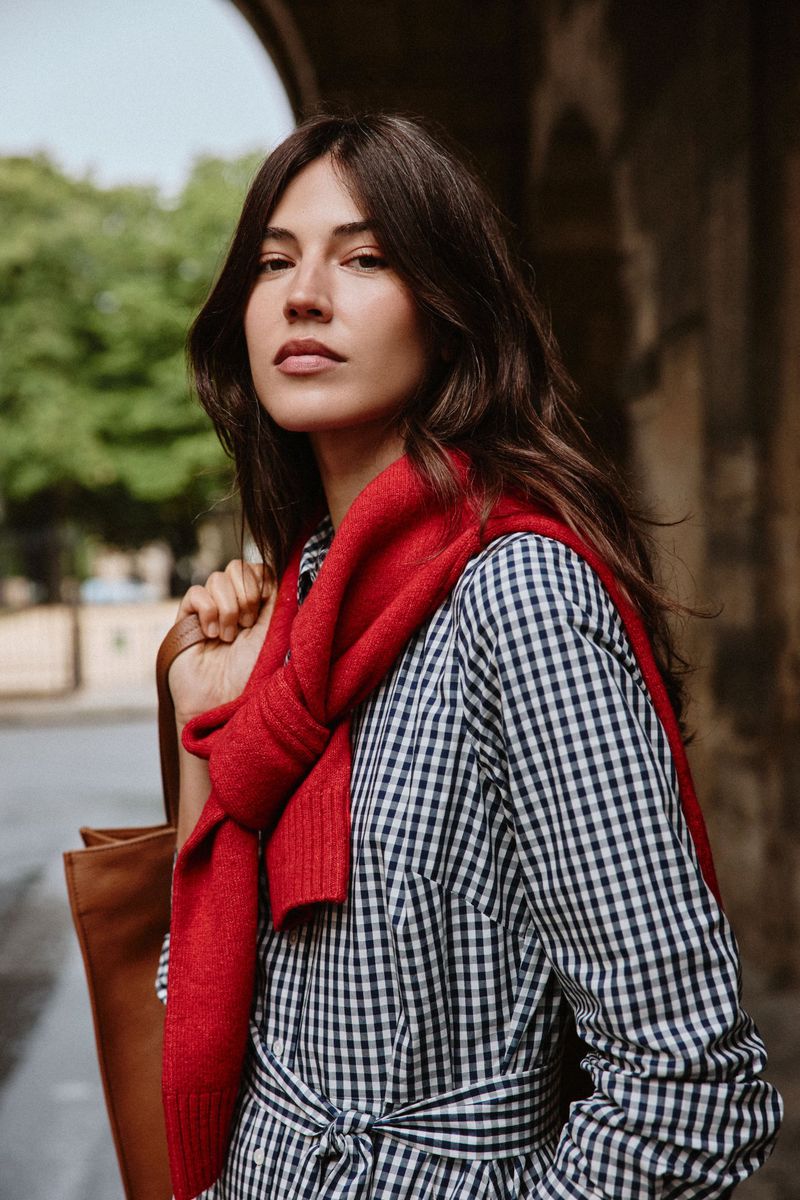 A brunette woman in a gingham dress and red sweater poses outdoors against a stone wall carrying a brown leather tote bag.