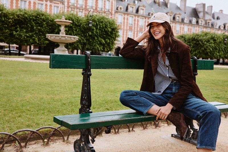 Smiling woman in jeans and jacket sitting on a park bench near a fountain and buildings on a sunny day in a European city.
