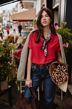 Model Marilhea Peillard in a red short-sleeve shirt, leopard scarf, and blue jeans holding flowers on a city street.
