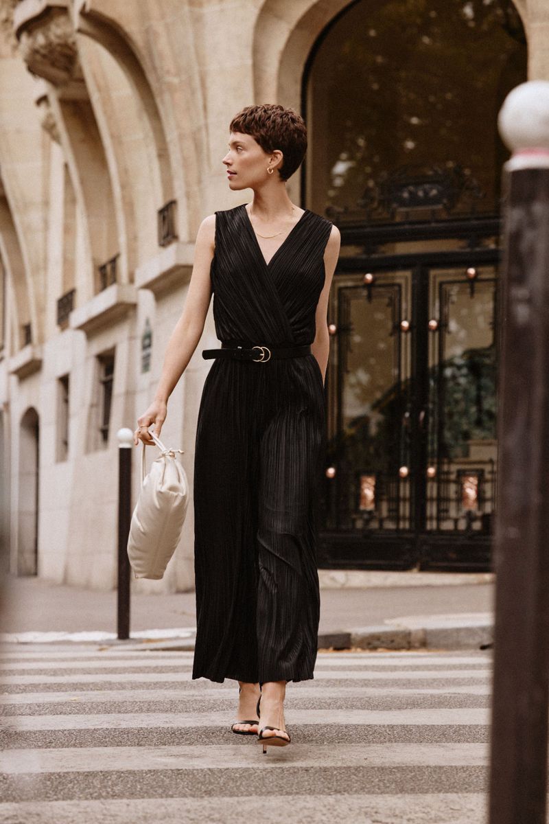 Model Loane Normand crosses a street wearing a pleated black sleeveless jumpsuit and holding a cream bag in a city setting.