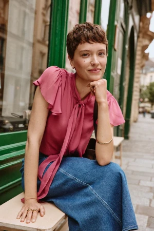 Young woman with short brown hair poses while seated outdoors wearing a pink blouse, blue denim skirt, and gold jewelry.