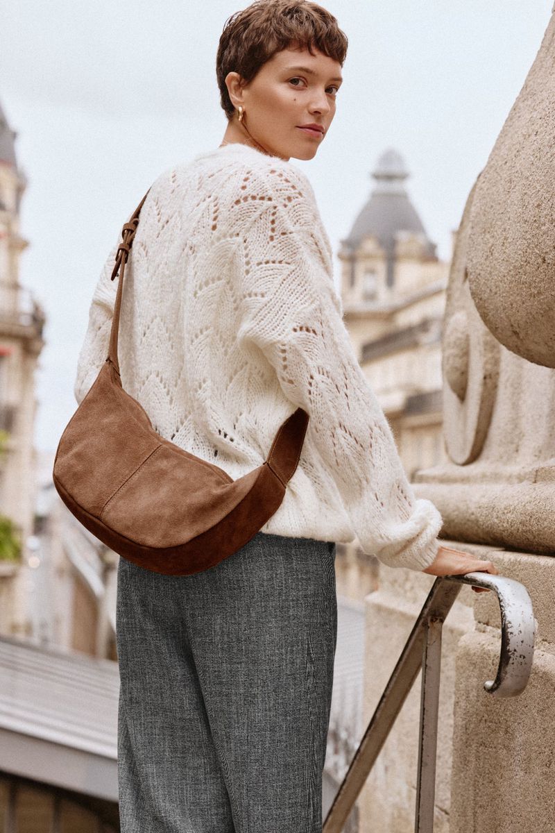 A woman in a cream sweater and grey trousers wears a brown crescent-shaped suede bag in an outdoor urban environment with a stone balustrade.