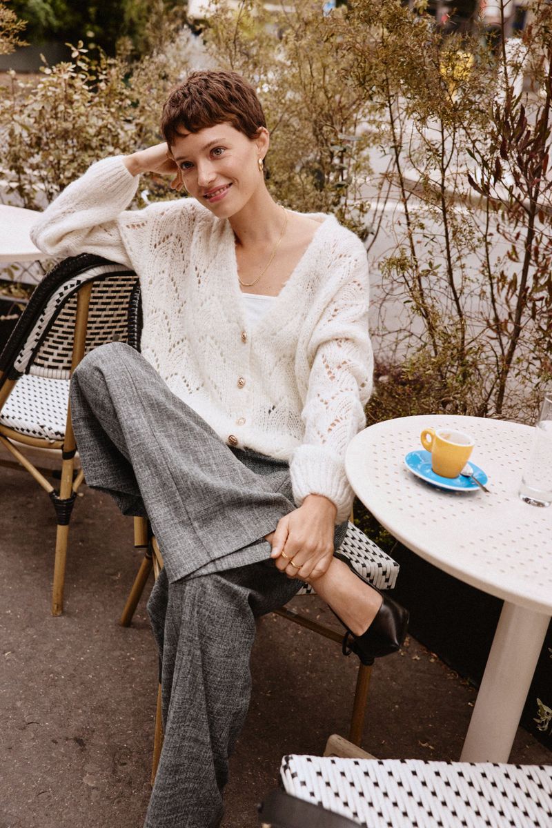 A serene outdoor cafe scene features a smiling woman in a white cardigan and grey trousers, seated next to a round table with a coffee cup.