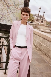 Model with short brown hair wearing a pink pantsuit and white t-shirt standing outdoors near a street map board.