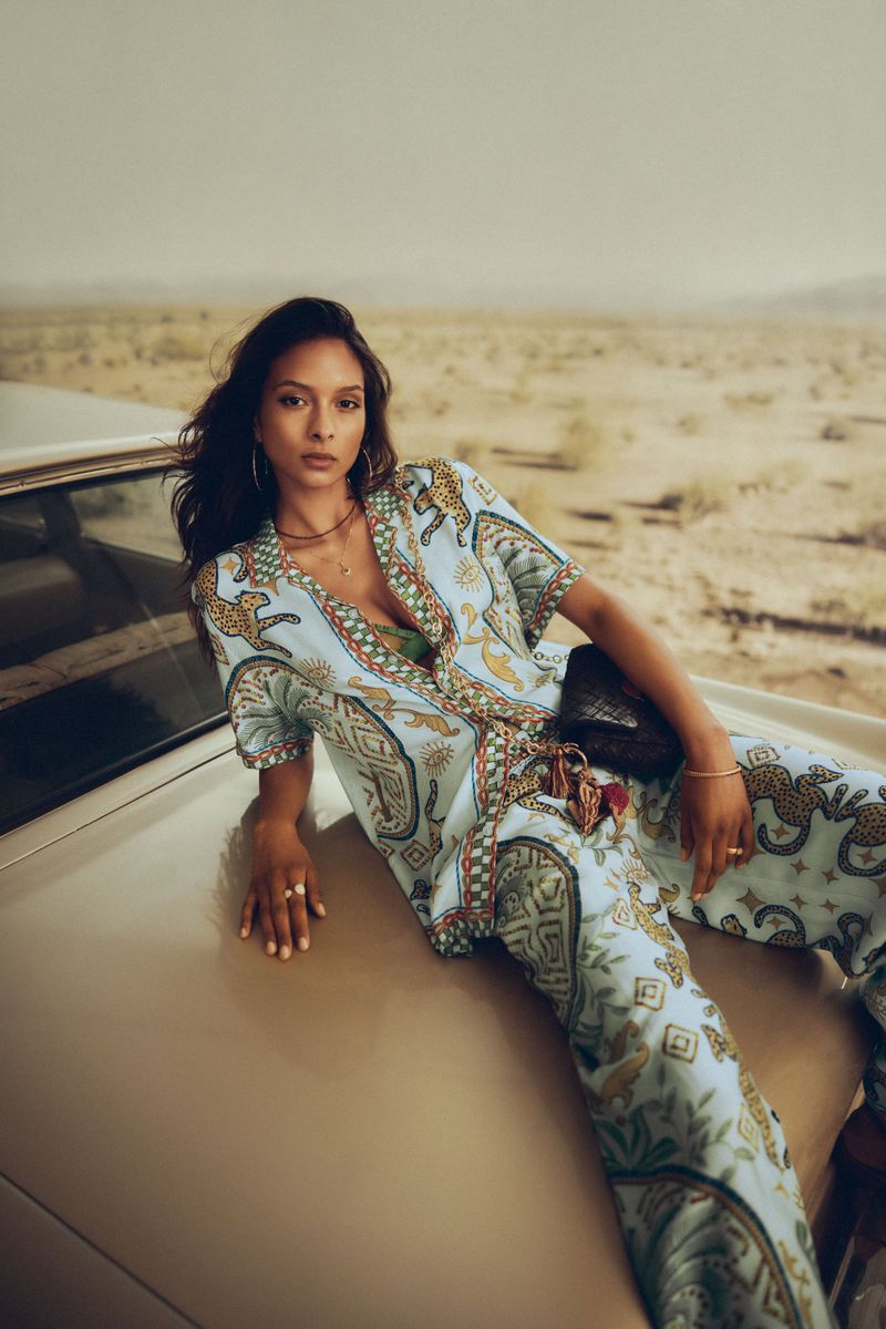 A woman in patterned pajama-style clothing poses on the hood of a vintage car in a desert landscape under an overcast sky.