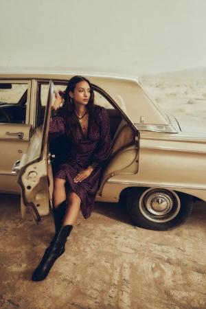 A woman in a patterned dress sits in the open doorway of a classic car in a desert landscape wearing black leather boots.