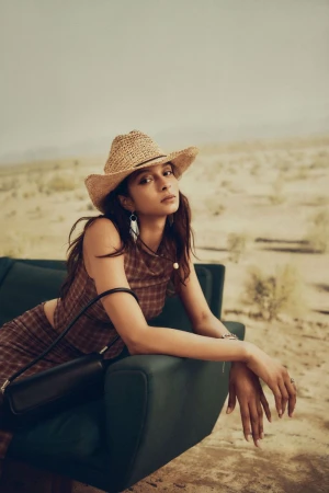 Woman in a plaid outfit and straw cowboy hat sits on a green couch outdoors in desert with dark shoulder bag and neutral color grading.