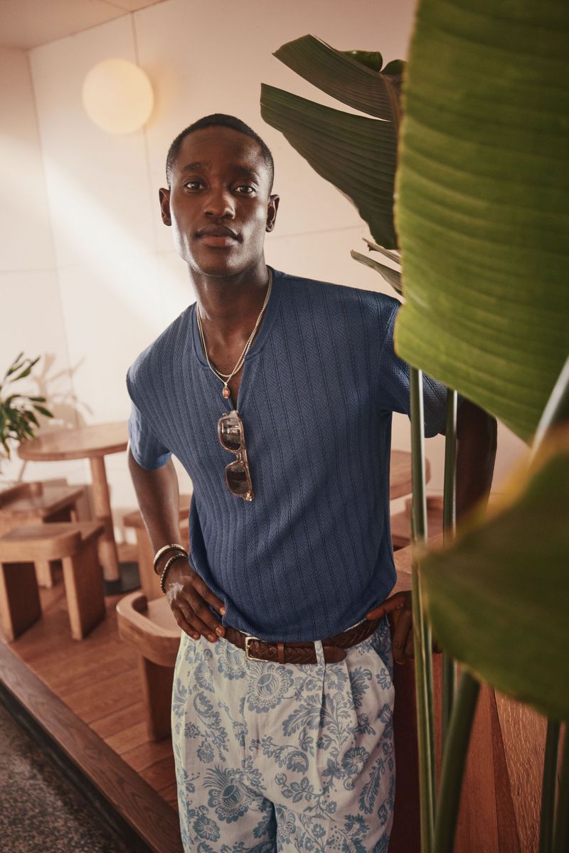Stylish dark-skinned man in blue shirt and patterned shorts stands indoors with plant and wooden furniture in the background.