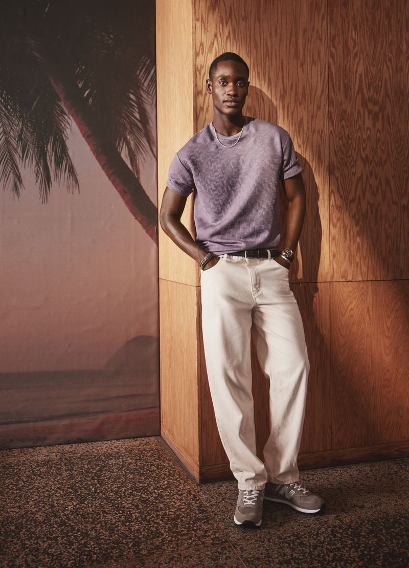 An adult African-descent man poses in a studio-like indoor setting, wearing a mauve tee, light-colored jeans, and grey sneakers, standing with his hands in his pockets.