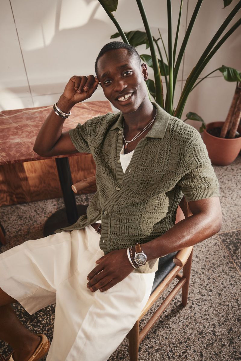 Smiling adult male in a green knitted shirt, off-white shorts, sitting in a chair near a wooden table and indoor plants.
