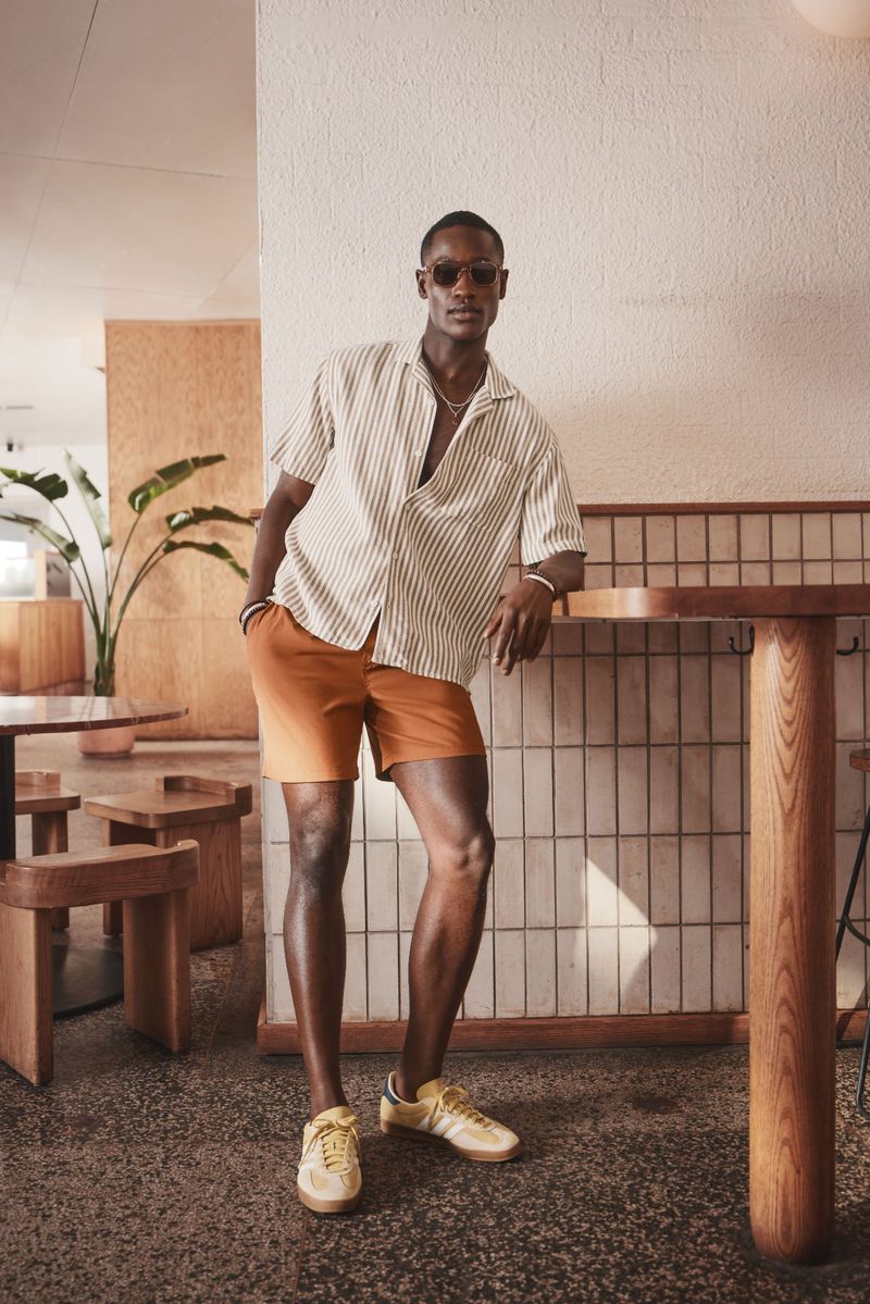 Attractive man in a striped shirt and shorts stands near a bar table in a bright, airy indoor setting with natural light.