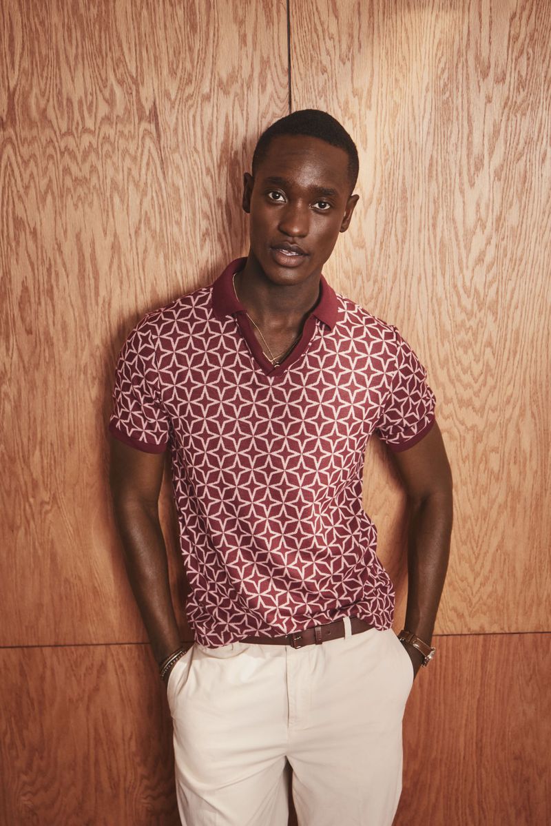 Portrait of a dark-skinned man with hands in pockets wearing a patterned polo shirt and light pants standing against a wood background.
