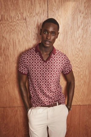 Portrait of a dark-skinned man with hands in pockets wearing a patterned polo shirt and light pants standing against a wood background.