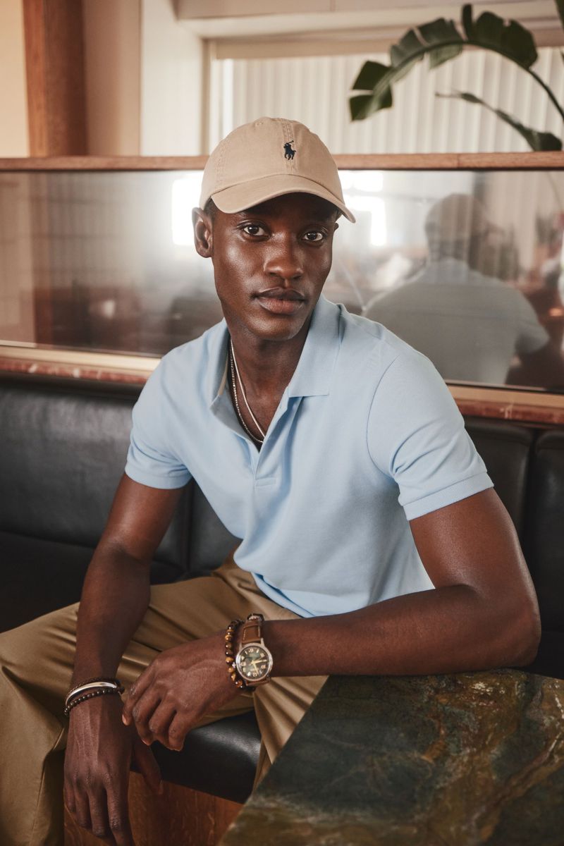 Black man in tan cap and blue polo shirt sitting indoors. He wears a watch and bracelets, with green plants visible in background.
