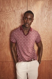 Model Charles Oduro wearing a burgundy patterned polo shirt and white trousers against a wood panel wall