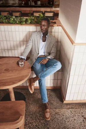 Model Charles Oduro seated in a grey blazer and blue jeans at a café for Simons Canada menswear editorial by D. Picard.