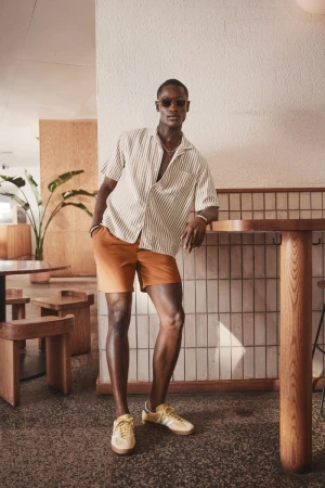 Model Charles Oduro leaning against a wooden cafe counter wearing a striped short-sleeve shirt, orange shorts, and yellow sneakers.