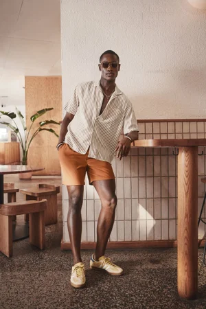 Model Charles Oduro leaning against a wooden cafe counter wearing a striped short-sleeve shirt, orange shorts, and yellow sneakers.