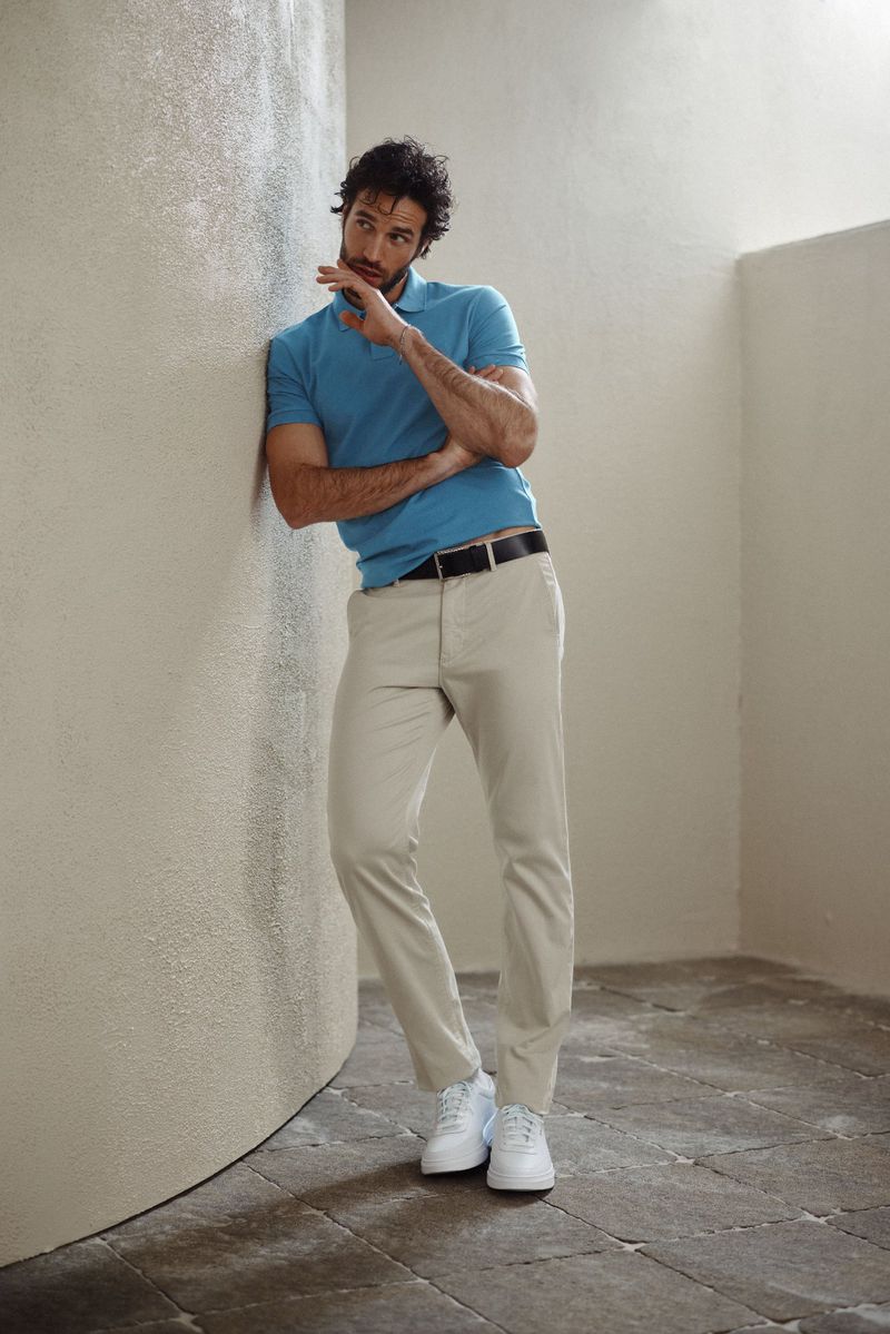 Man in polo and chinos leaning against a wall. He is wearing white sneakers. Soft light illuminates the corner.