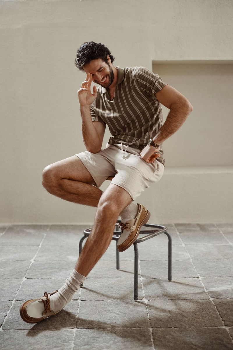 Man wearing striped polo shirt and beige shorts sitting on a metal stool against a neutral wall