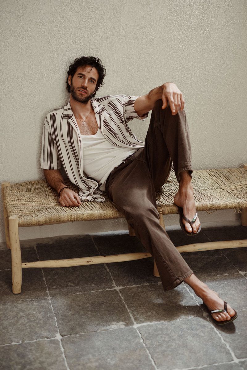 Dark-haired man poses on a bench in casual summer fashion, wearing a striped shirt and dark pants, set against a neutral wall.