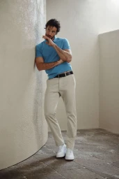 Model Paul Ferrari leaning against a neutral stucco wall wearing a blue polo shirt, beige chinos, and white sneakers.
