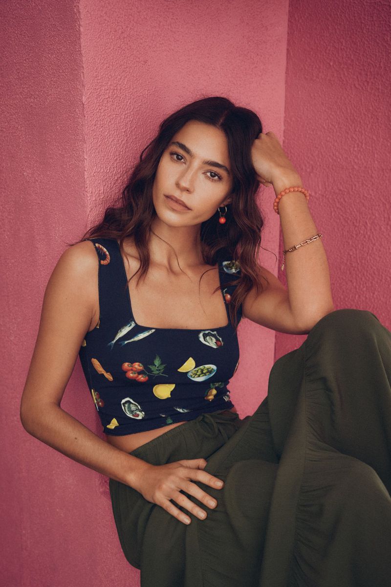 A young woman with wavy hair poses in front of a pink wall, wearing a navy crop top with food illustrations and olive-green pants.