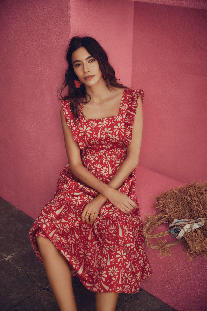 Woman in a red floral dress sits against a pink wall with a straw bag. Shoulder-length wavy hair and red earrings complete her look.
