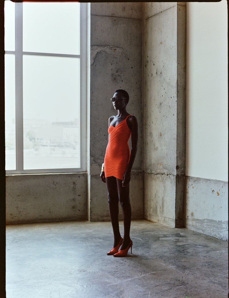Woman in an orange dress and sunglasses standing in a minimalist room with concrete walls and a large window, fashion photography.