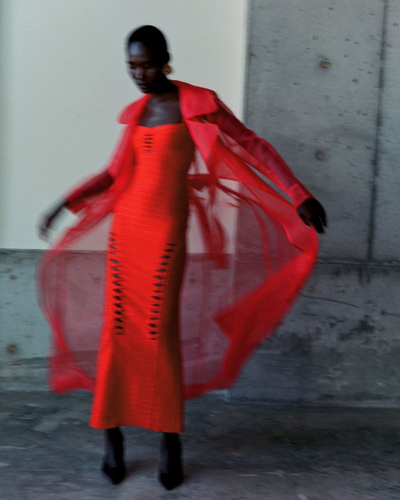 Woman poses in an orange dress and sheer orange coat against a concrete wall for a fashion editorial, shot with motion blur.