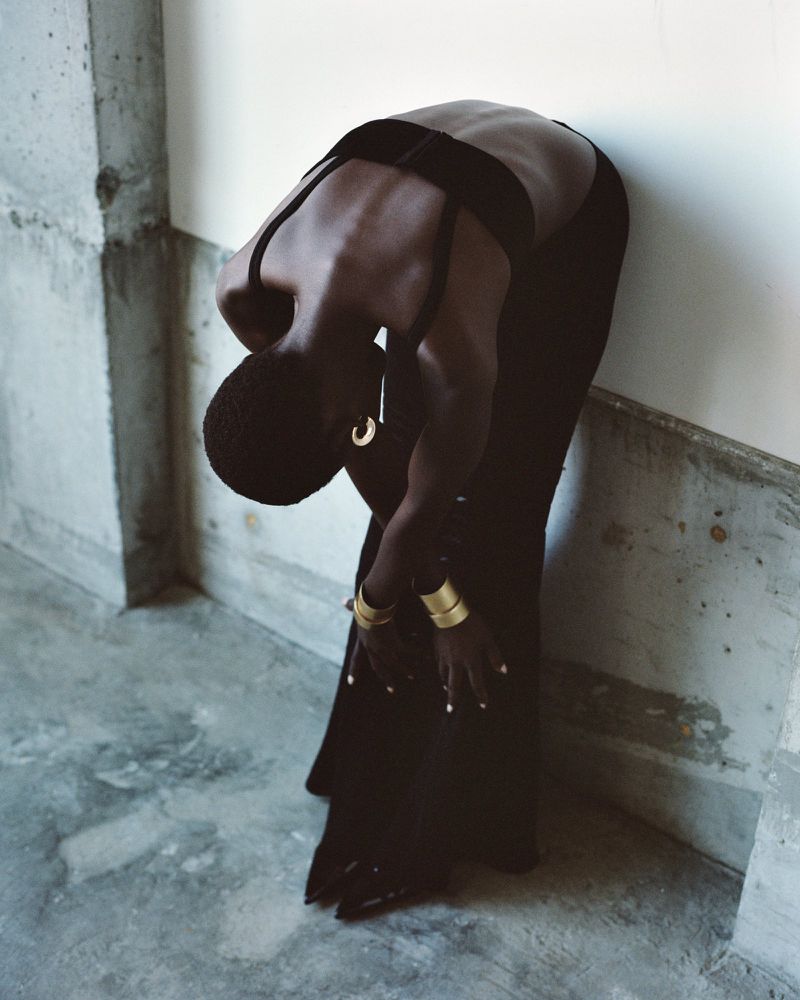 Woman in black crop top and pants leans against a concrete wall, showcasing gold bracelets and earrings in a minimalist fashion setting.