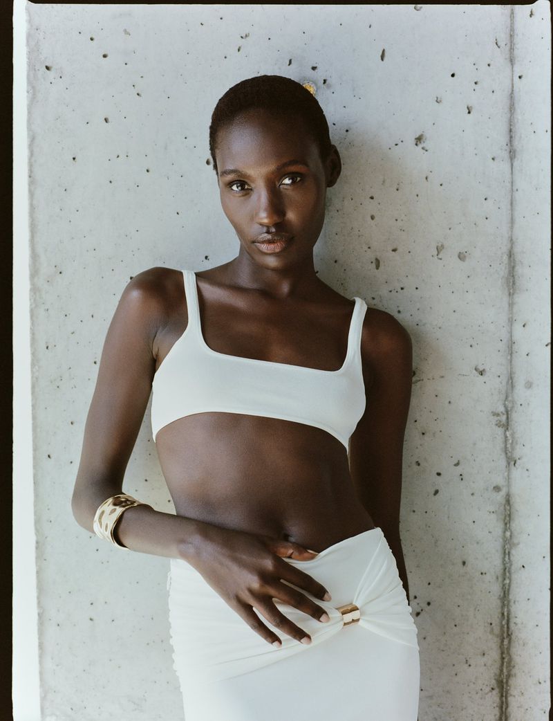 Close-up of a black woman in a white two-piece outfit against a concrete wall, accessorized with a gold cuff bracelet. 