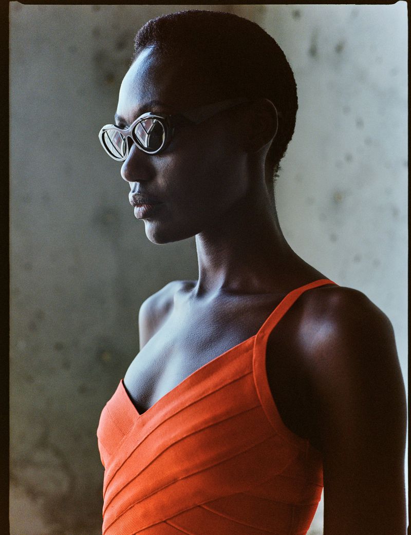 Woman in orange dress and stylish sunglasses poses in a fashion shot with soft lighting and concrete background; beauty, fashion, portrait.