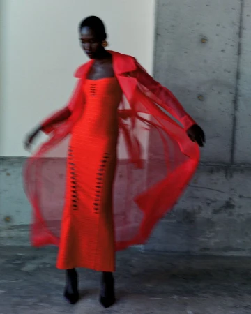 Model in a bright red sheer trench coat and matching dress against a concrete wall