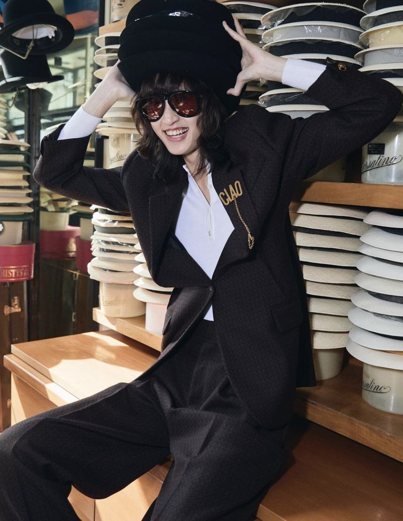 A stylish young woman in a black suit and hat sits surrounded by rows of hats, showcasing a vintage fashion look with dark sunglasses.