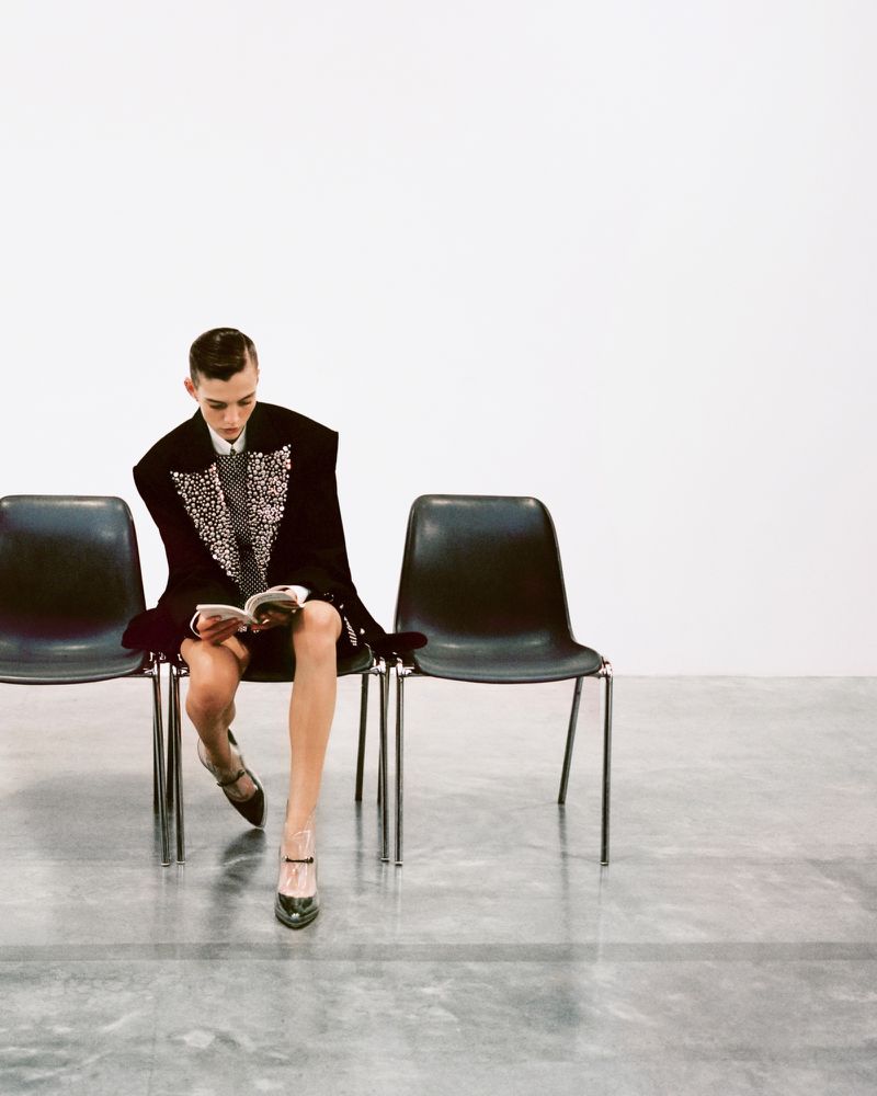 Stylish woman in embellished blazer and heels sits reading on one of three chairs in a minimalist white photo studio setting.