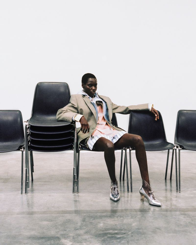 An African-American woman poses while sitting in a chair between stacks of chairs wearing a blazer, patterned dress, and silver heels indoors.