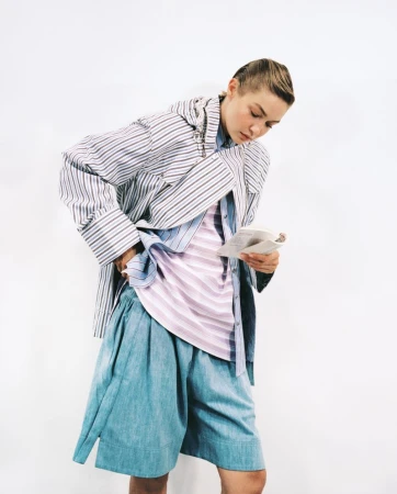 Woman in a striped shirt and blue shorts reads a newspaper in a brightly lit studio setting, captured in a fashion editorial shot.
