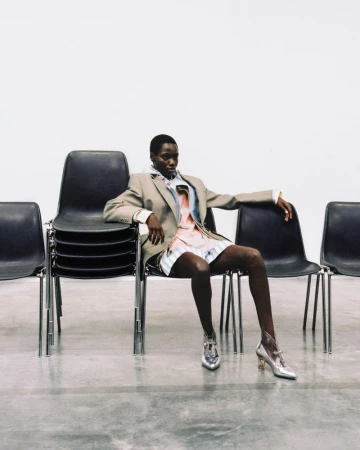 Model seated on a black chair wearing a beige blazer over a layered striped shirt and shiny silver shoes against a plain white background