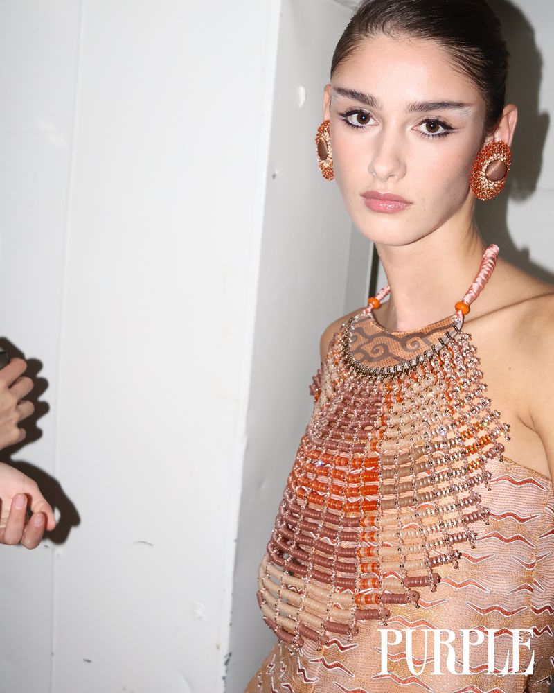 A poised fashion model stands indoors wearing a beaded halter top and statement earrings against a white wall, showcasing a unique style.