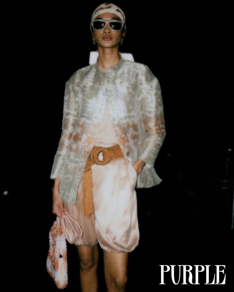 A model poses in an ethereal outfit, wearing sunglasses and a headscarf, against a dark backdrop. The outfit includes a sheer jacket.