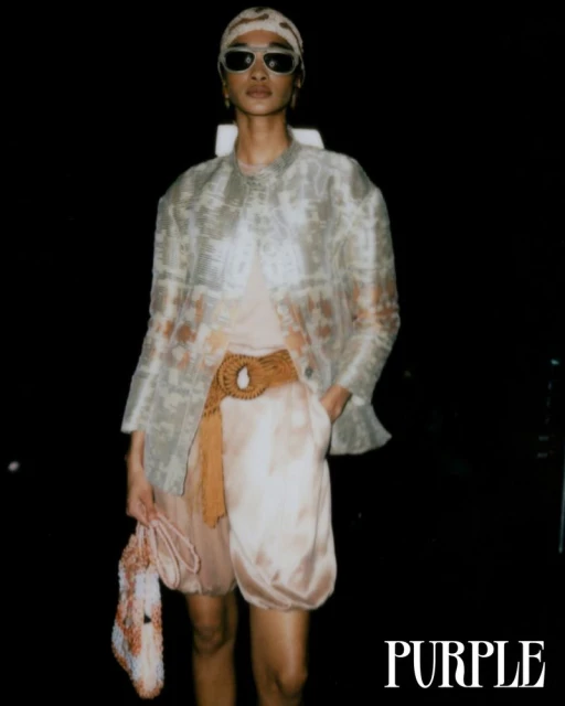 A model poses in an ethereal outfit, wearing sunglasses and a headscarf, against a dark backdrop. The outfit includes a sheer jacket.