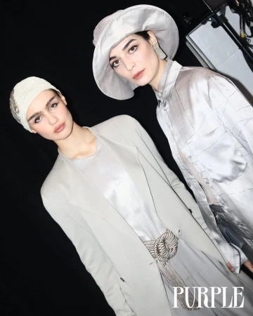 Two fashion models in matching grey outfits and hats pose indoors against a black backdrop, styled for an editorial or runway setting.