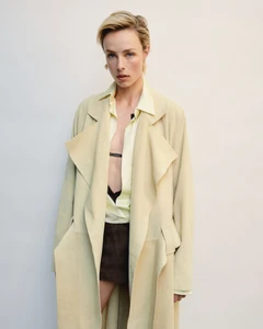 Model wearing a pale yellow trench coat and open collared shirt against a plain seamless white backdrop