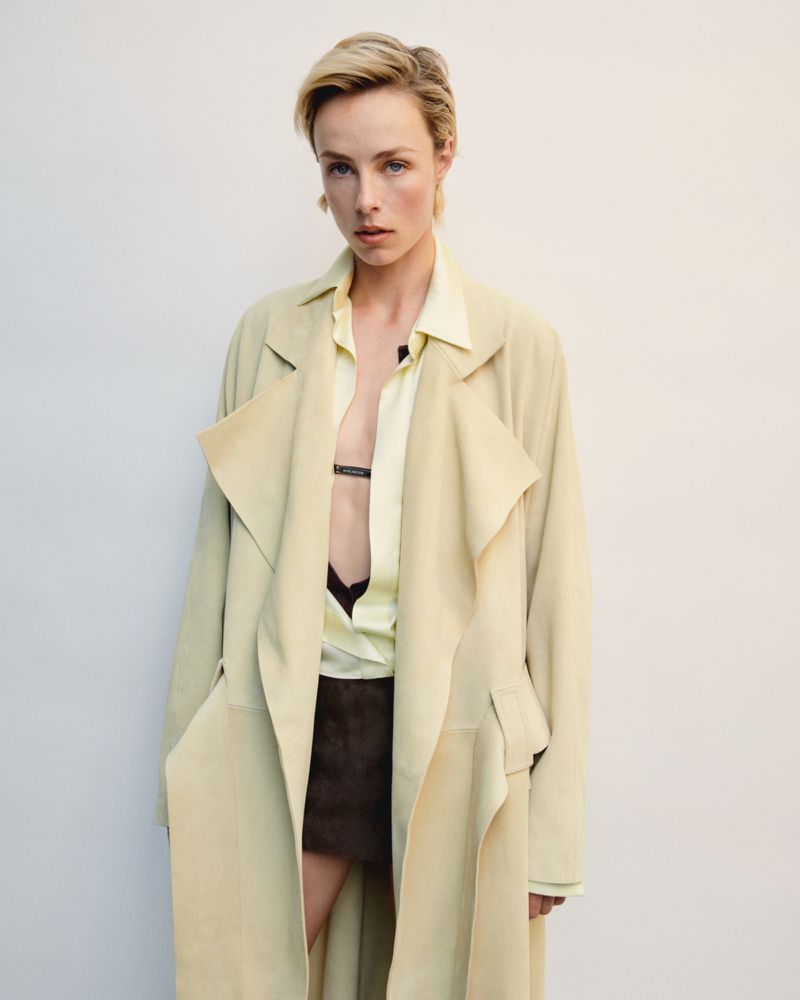 A fair-skinned female model with a blond pixie cut poses in a light yellow button-down shirt, brown miniskirt, and a long, light yellow coat.