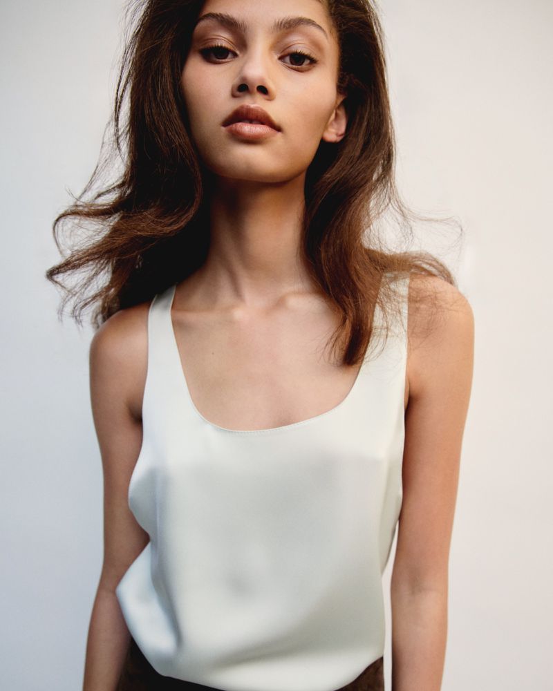 Portrait of a young adult woman with long brunette hair in a sleeveless, light-green top, gazing towards the camera in a studio setting.