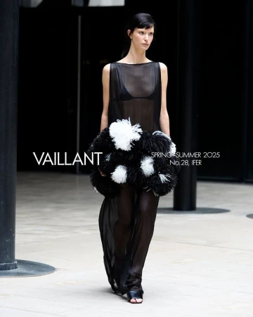 Fashion model on runway wearing sheer black dress with black and white pom-pom embellishment. Runway show set indoors with dark background.
